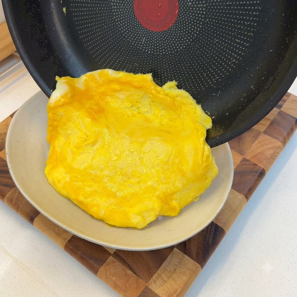 Ein weich gekochtes Omelett gleitet aus einer Bratpfanne und drapiert sich über eine Portion weißen Reis.