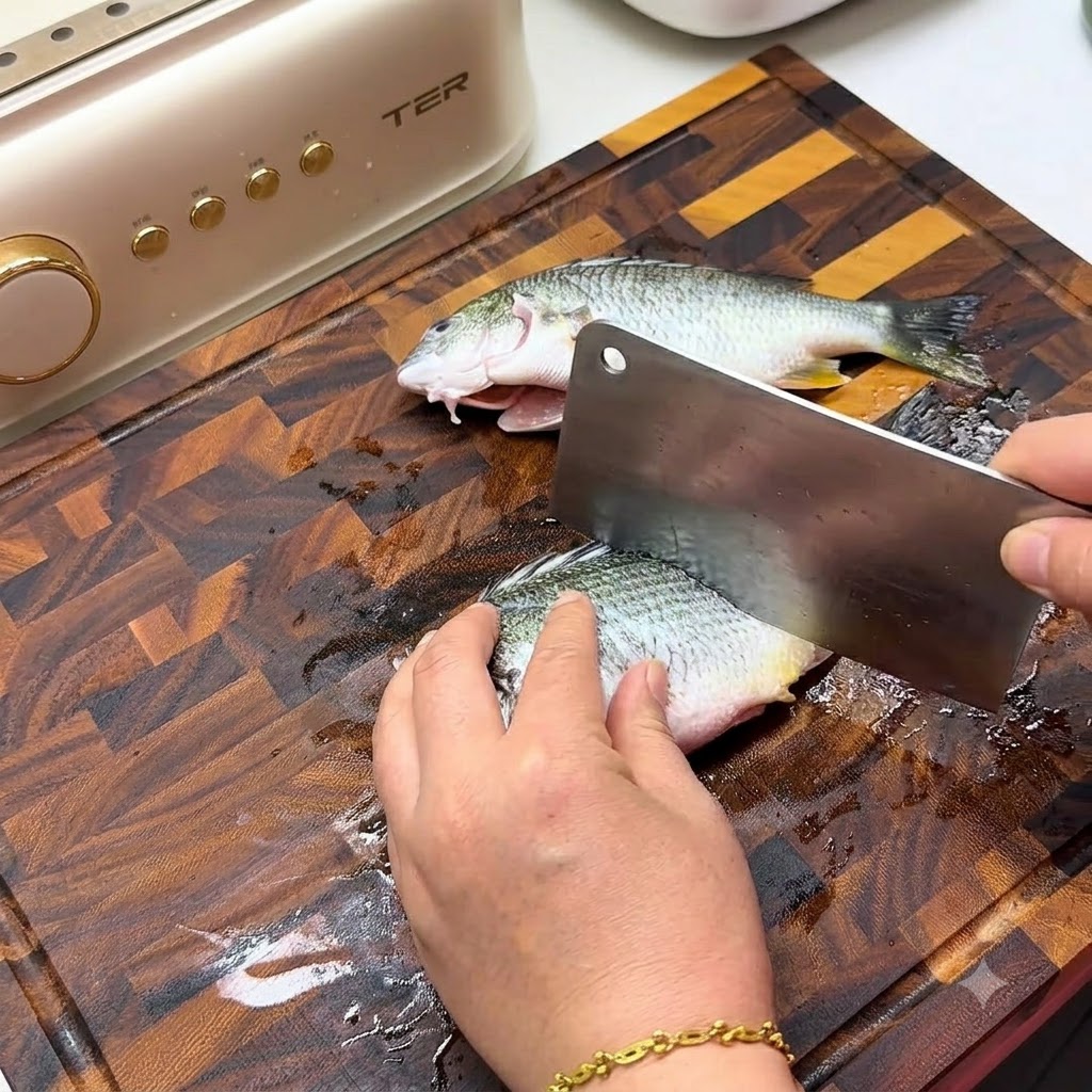 Eine Hand benutzt ein großes Metallsbeil, um diagonale Schnitte auf der Seite eines frischen Fisches auf einem Holzschneidebrett zu machen.