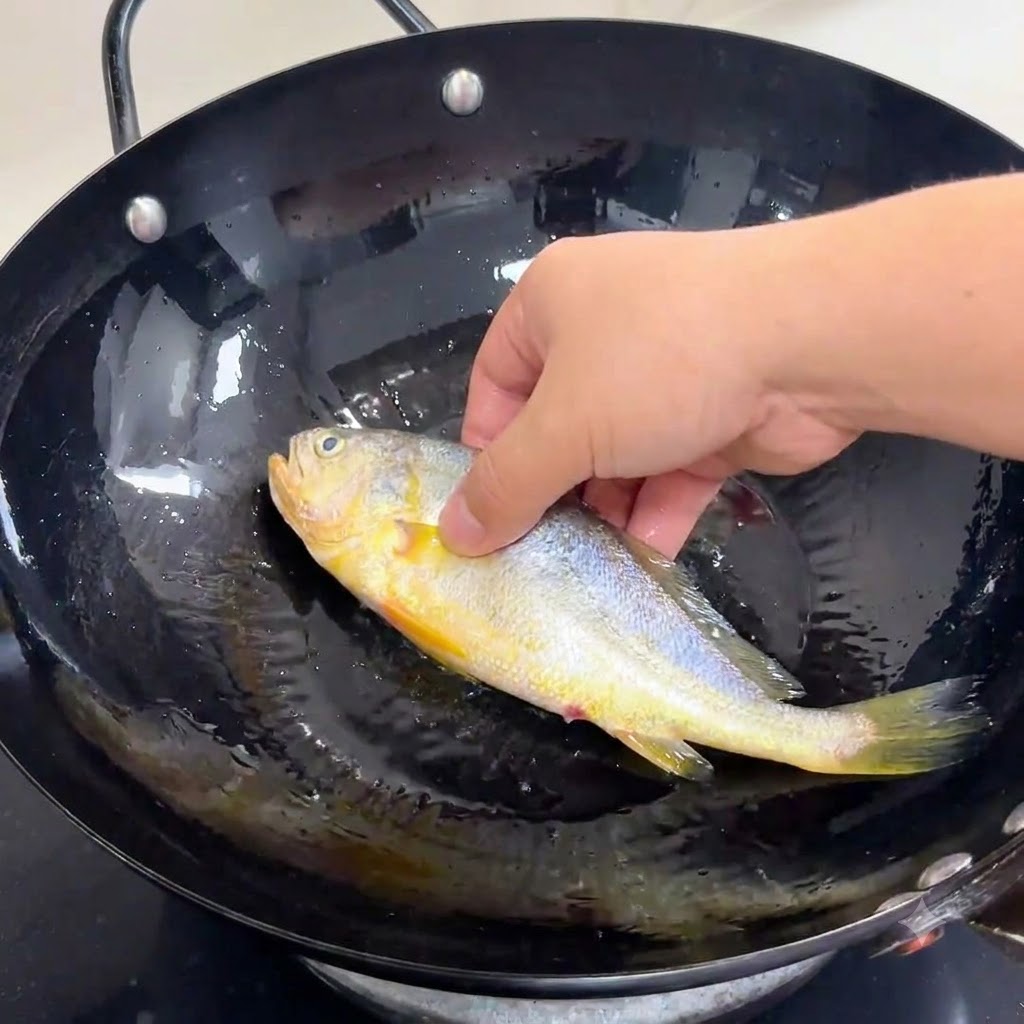 Eine Hand legt einen gewürzten ganzen Fisch vorsichtig in einen schwarzen Wok mit erhitztem Speiseöl.