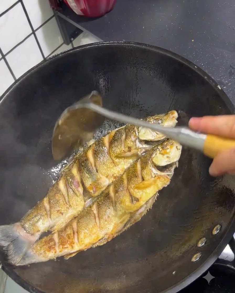 Zwei Gelbschwanzfische braten in einem Wok mit goldbrauner Haut, gewendet mit einem Pfannenwender.