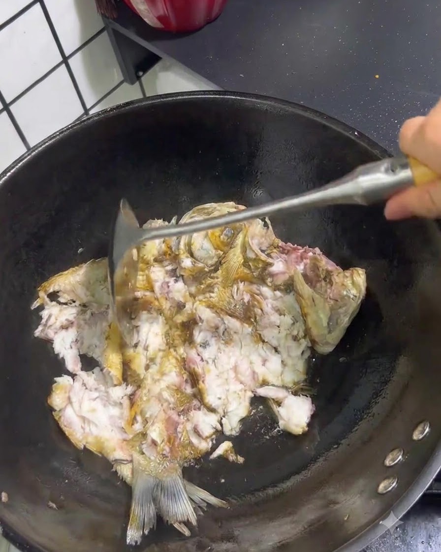 Gebratener Fisch wird mit einem Metallpfannenwender in einem Wok auseinandergebrochen und zerdrückt.