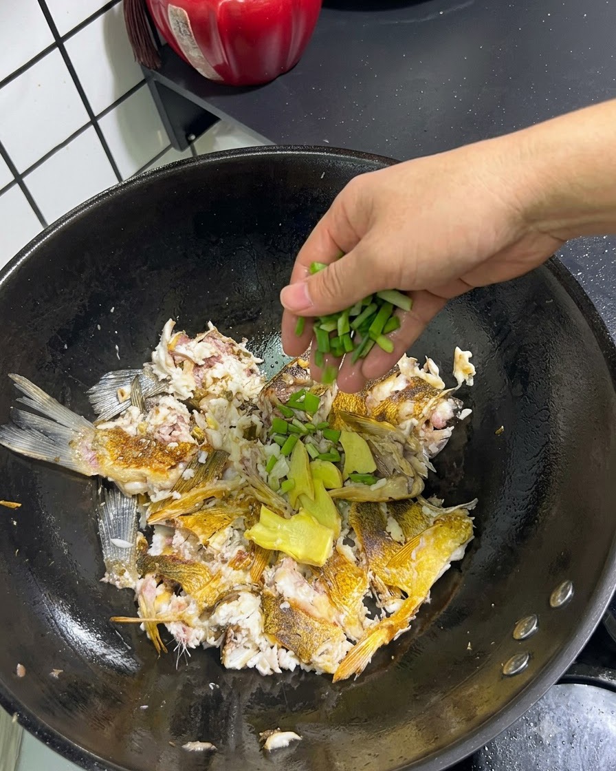 Frische Frühlingszwiebeln und in Scheiben geschnittener Ingwer werden von Hand über den zerdrückten Fisch im Wok gegeben.