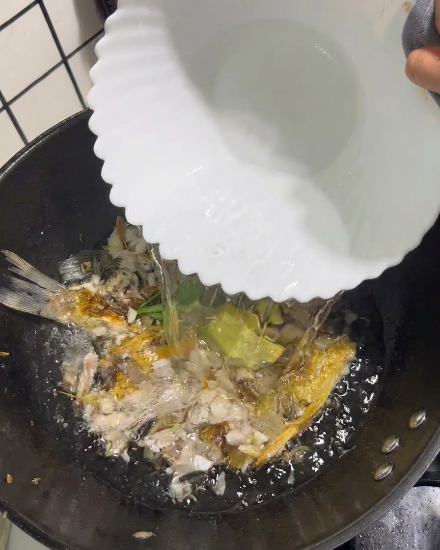 Klares kochendes Wasser wird aus einer weißen Schüssel in einen Wok mit zerdrücktem Fisch und Aromaten gegossen.