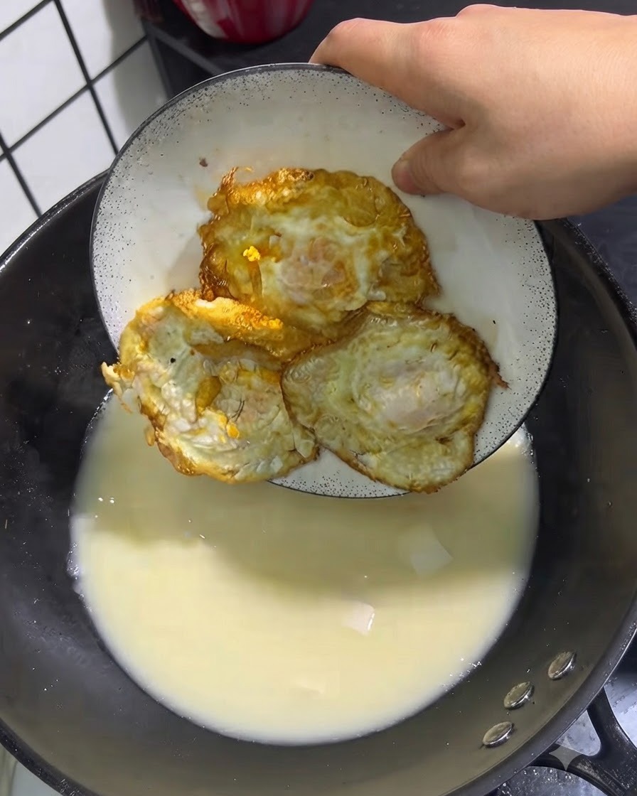 Drei goldgebratene Eier werden von einem weißen Teller in einen Topf mit köchelnder Suppe gelegt.