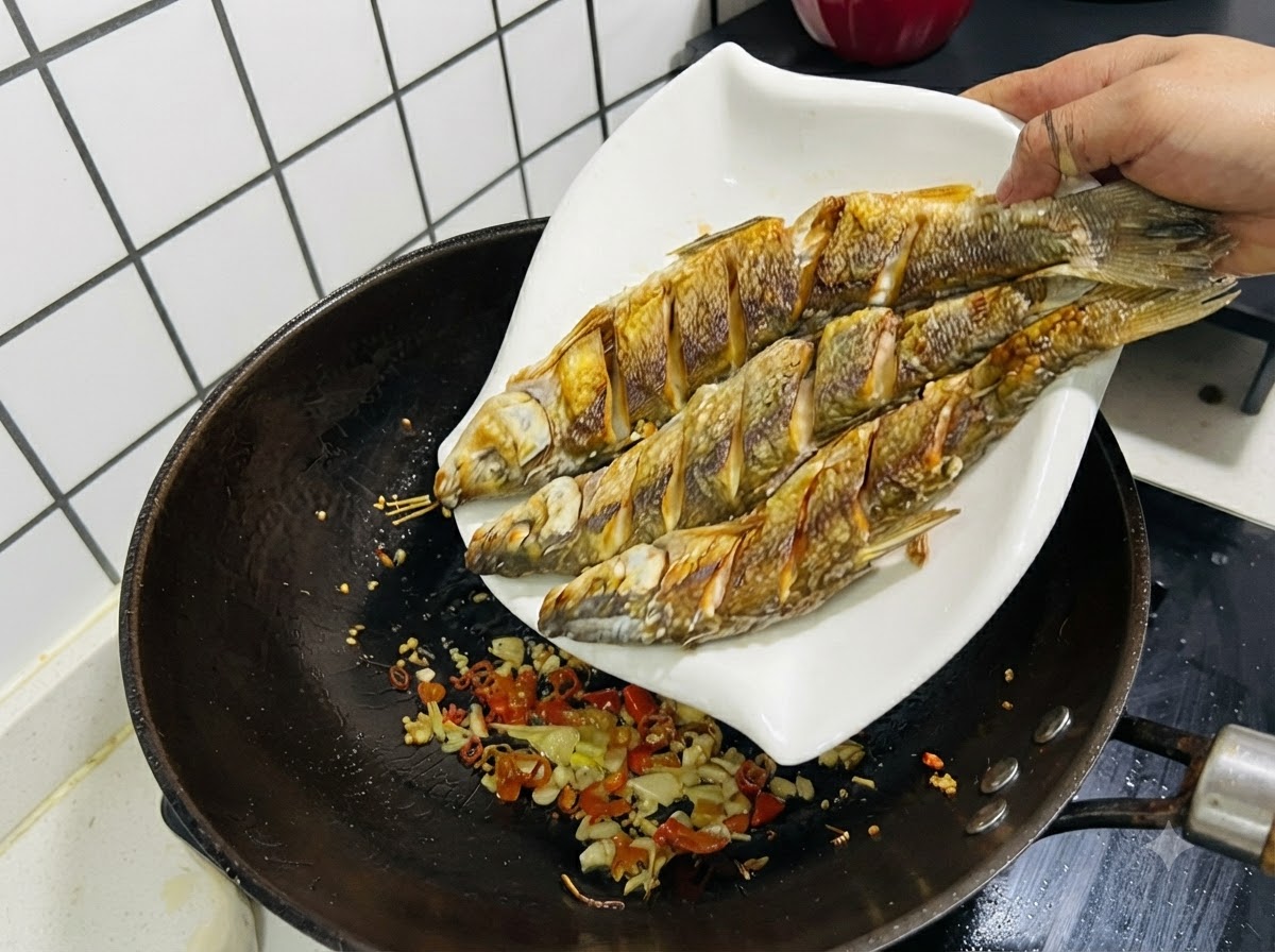Drei frittierte Fische werden von einem weißen Teller zurück in einen Wok mit sautiertem Knoblauch, Ingwer und Chilischoten geschoben.