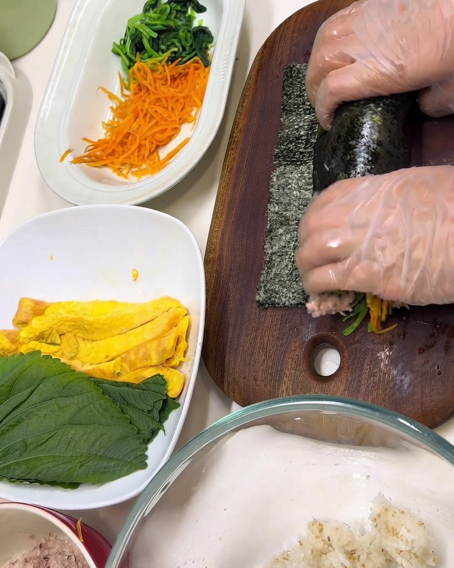 Hände mit transparenten Handschuhen wickeln Zutaten auf einem Holzschneidebrett in ein Nori-Blatt ein.