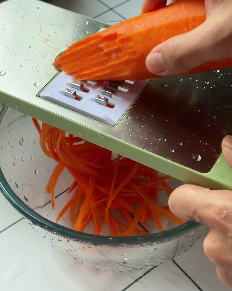 Eine Hand hält eine geschälte Karotte an eine grün gerahmte Reibe und raspelt sie in eine Glas schüssel.