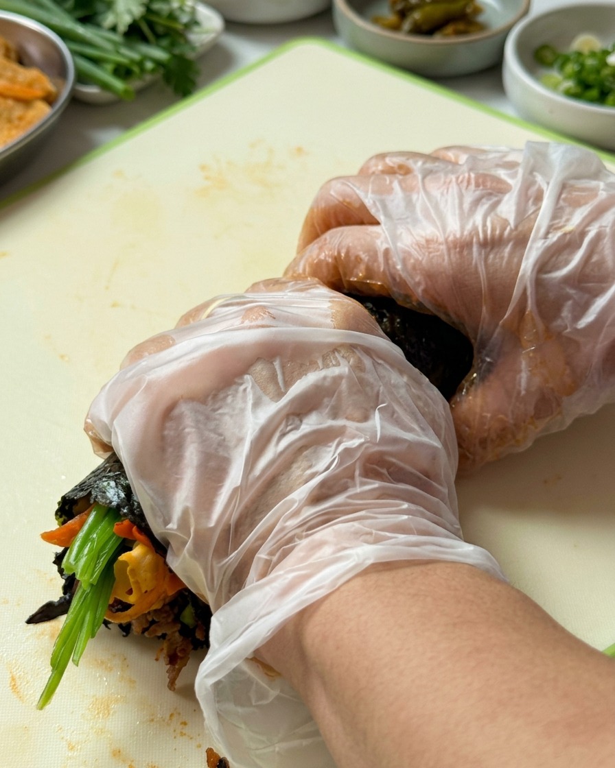 Hände mit Handschuhen rollen das Gimbap fest von der Unterkante nach oben auf.