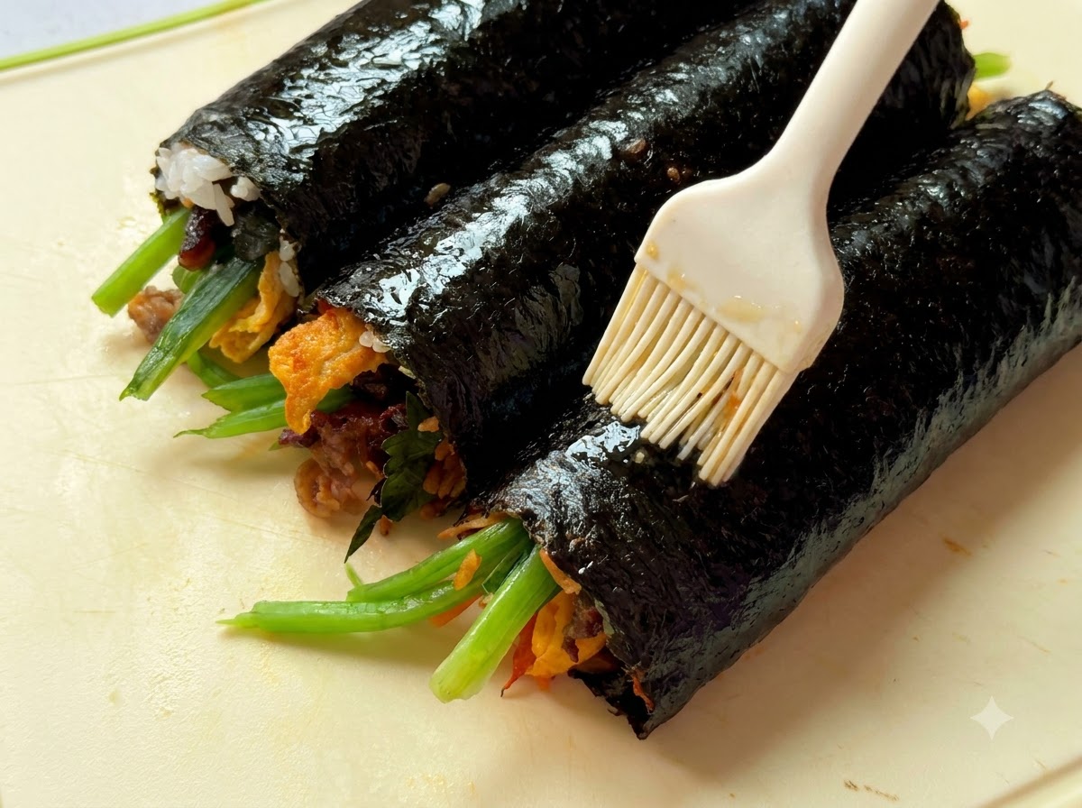 Nahaufnahme von Sesamöl, das mit einem Pinsel auf drei gerollte Gimbap-Rollen auf einem Schneidebrett aufgetragen wird.