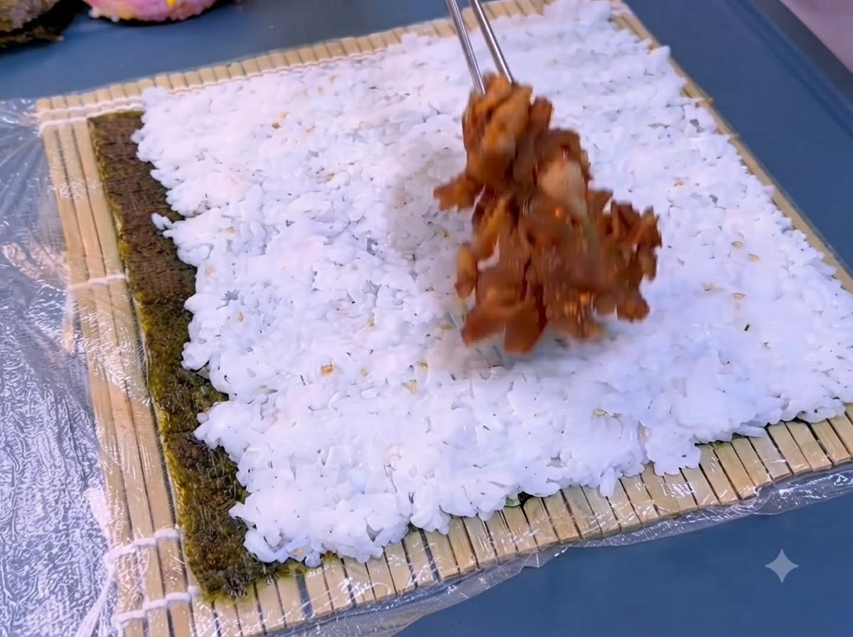Ein dunkles Algenblatt auf einer Bambusmatte, auf dem eine Schicht weißer, gekochter Reis verteilt wird.
