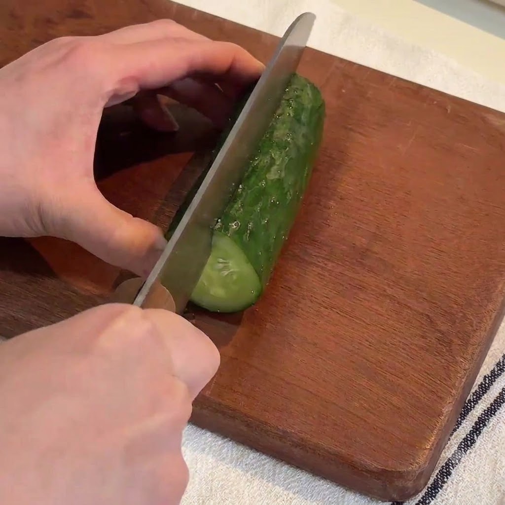 Hände schneiden mit einem großen Messer eine frische grüne Gurke auf einem Holzschneidebrett in lange Streifen.