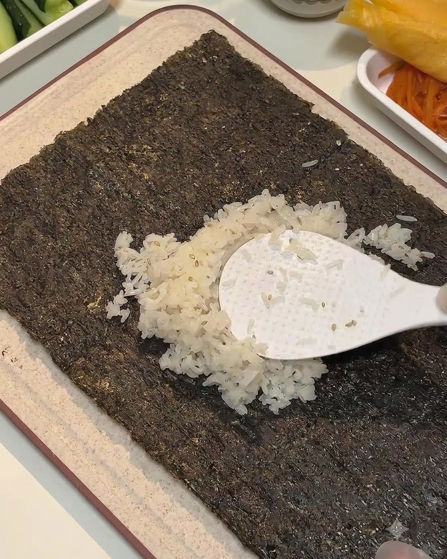 Ein weißer Reislöffel verteilt gewürzten Reis auf der Mitte eines Algenblattes.