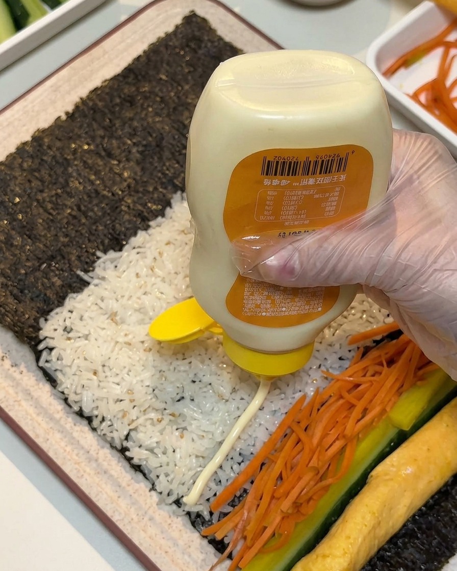 Eine behandschuhte Hand drückt dicke Linien weißer Mayonnaise aus einer Flasche über die ausgelegten Füllungen auf Seetang und Reis.
