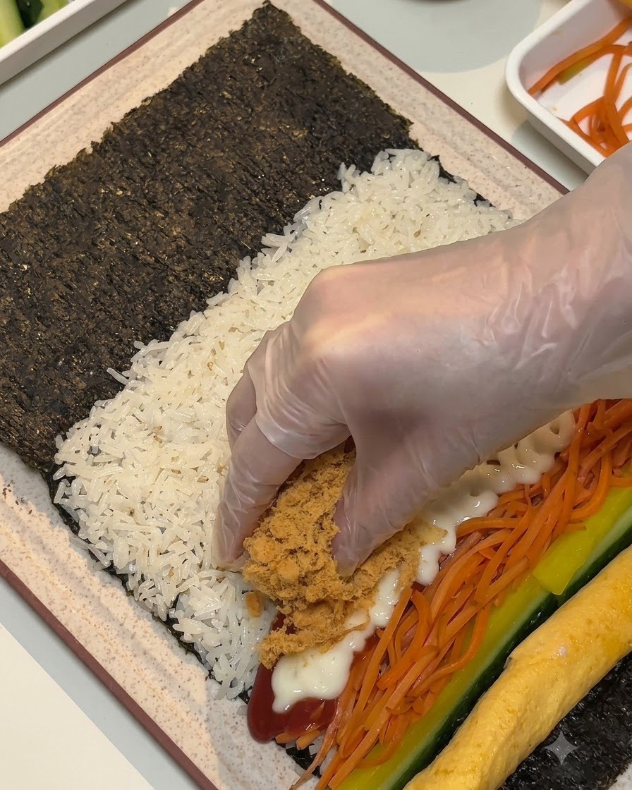 Eine behandschuhte Hand legt braune, fluffige Fleischwolle und Frühstücksfleischstreifen über die Füllungen auf das Algenblatt.
