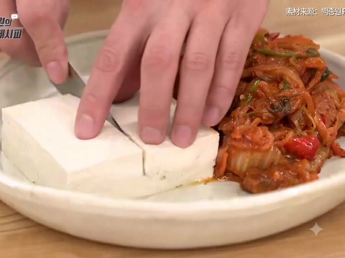 Eine Hand schneidet blanchierten Tofu sauber auf einem Teller neben einem Berg aus herzhaft gebratenem Kimchi und Schweinefleisch.