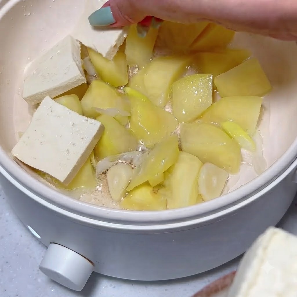 Tofuscheiben werden ordentlich über einer Schicht aus gekochten Kartoffelstücken und Knoblauch in einem weißen Topf angeordnet.