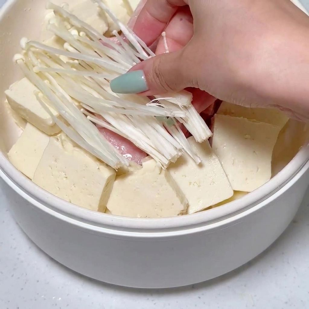 Enoki-Pilze werden zwischen angeordnetem Tofu und Frühstücksfleisch in den Topf gegeben.