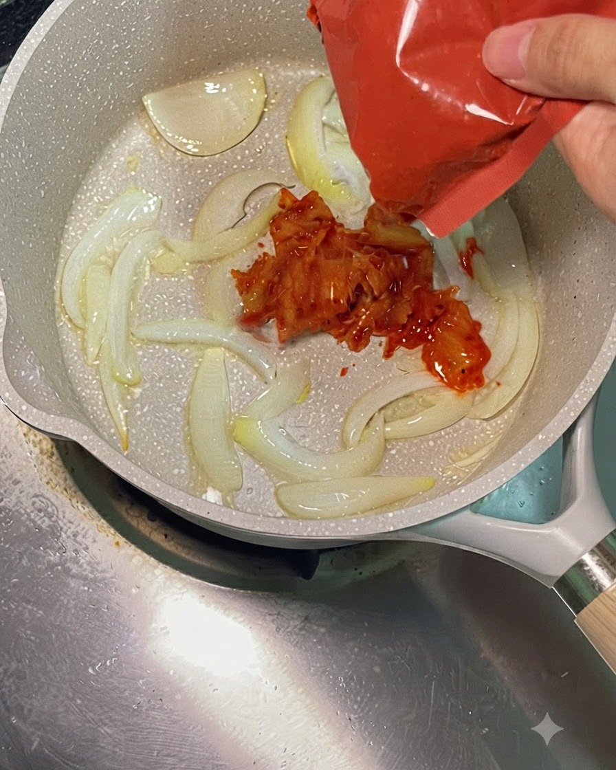 Leuchtend rotes, gehacktes Kimchi wird aus einer roten Verpackung in eine heiße Pfanne mit angebratenen Zwiebeln gegeben.