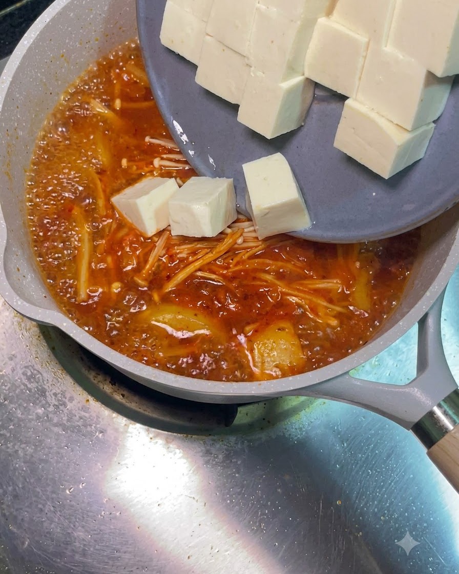 Weiße Tofuwürfel gleiten von einem grauen Teller in die brodelnde rote Suppe.