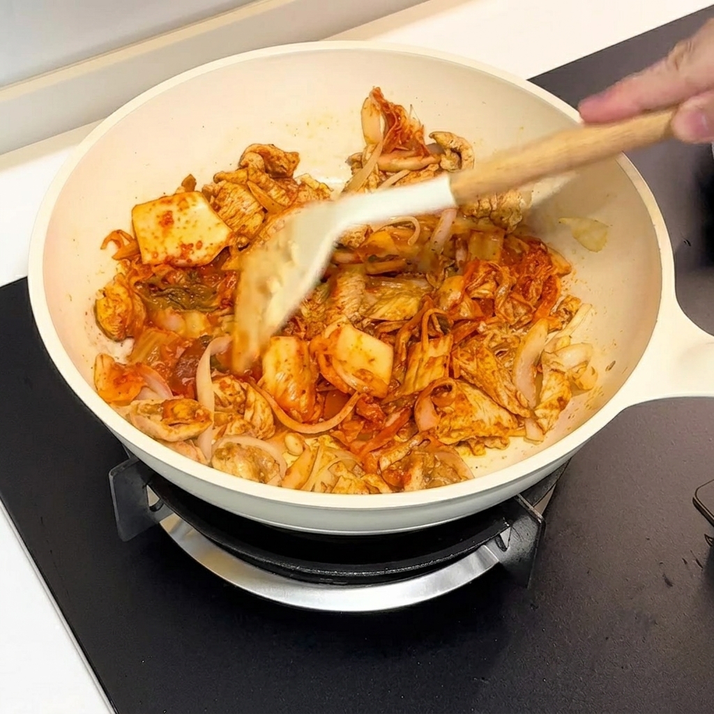 Kimchi und Hähnchen werden in einer weißen Pfanne mit einem Spatel zusammen angebraten.