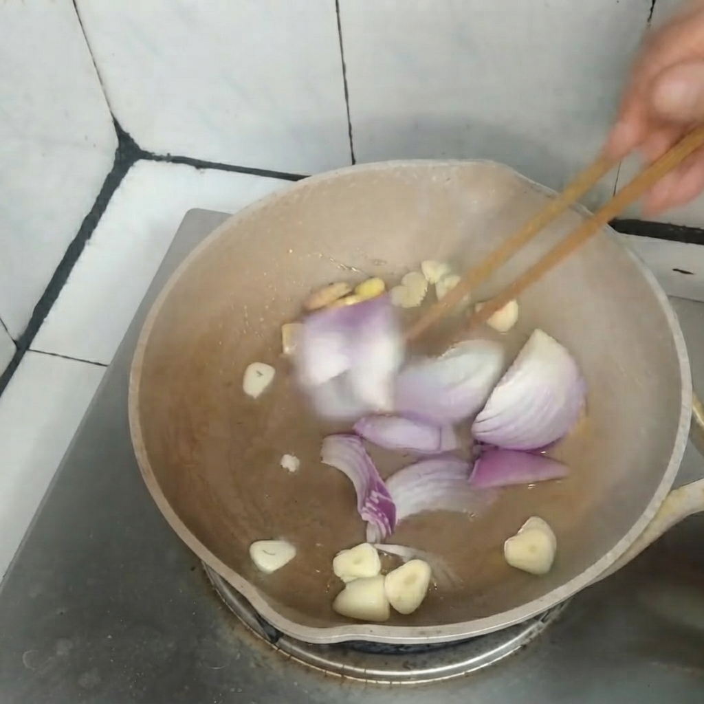Geschnittene rote Zwiebeln, Knoblauch und Ingwer werden mit Holzesstäbchen in einer Pfanne zusammen angebraten.