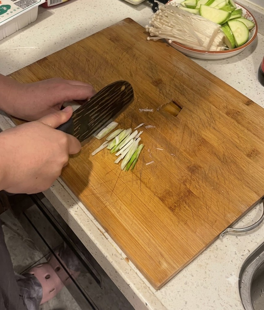 Hände schneiden mit einem Küchenmesser Frühlingszwiebelstangen auf einem Holzschneidebrett.