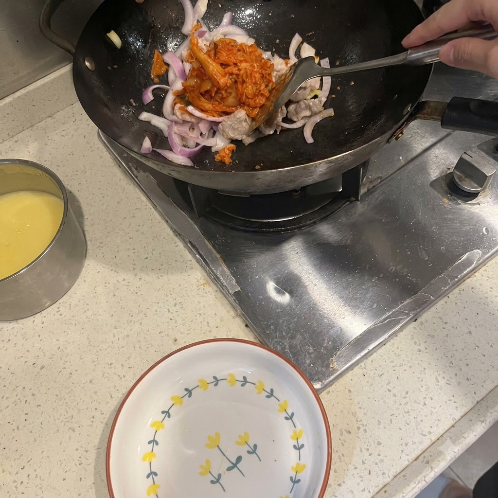 Ein Spatel gibt leuchtend rotes Kimchi in einen Wok mit angebratenem Schweinebauch und roten Zwiebeln.