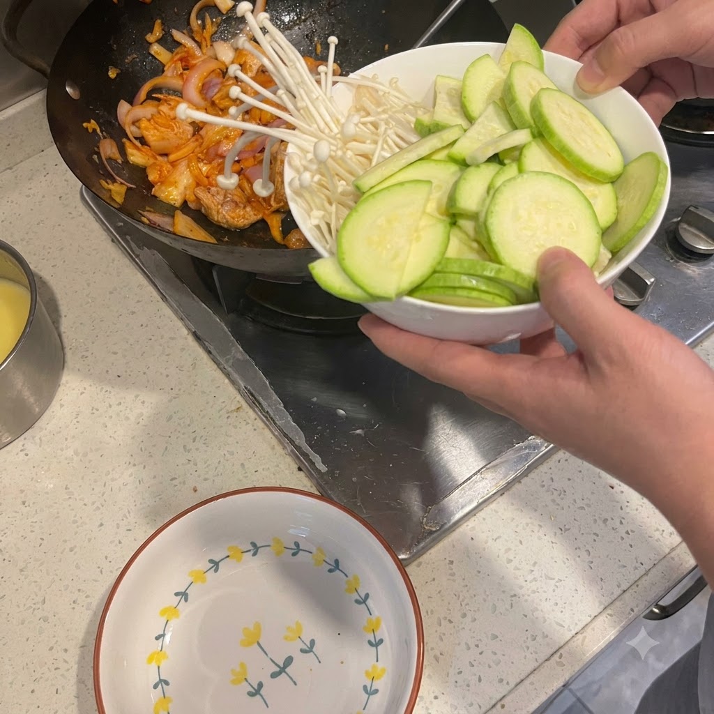 Geschnittene Zucchini und Enoki-Pilze werden aus einer Schüssel in einen Wok mit angebratenem Schweinefleisch und Zwiebeln geschüttet.