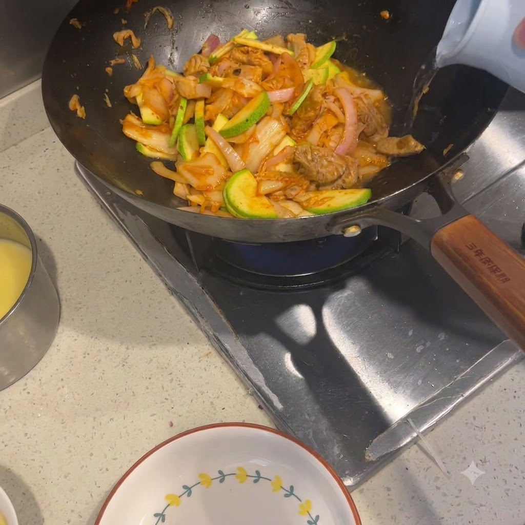 Heißes Wasser wird aus einem hellen Wasserkocher in einen Wok mit angebratenen Zutaten gegossen.