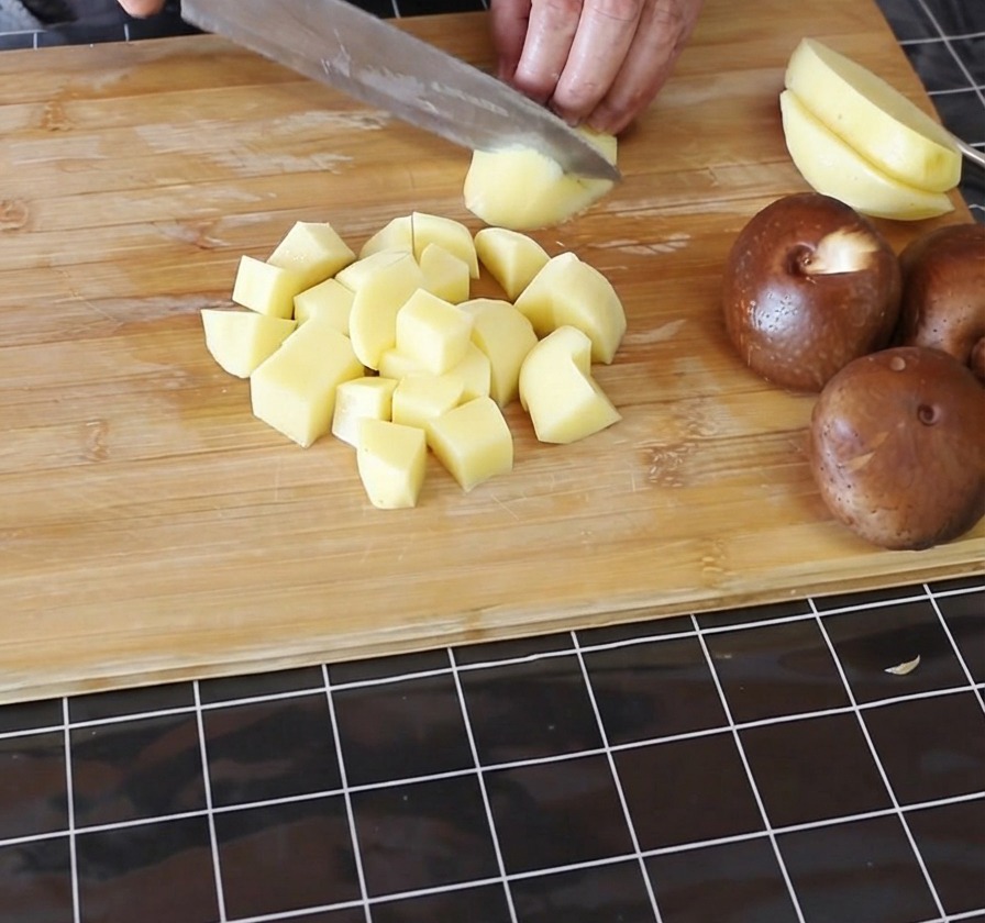Hände schneiden geschälte gelbe Kartoffeln auf einem Bambus-Schneidebrett in kleine Würfel.