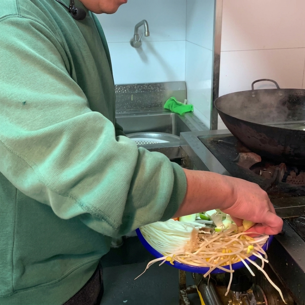 Eine Auswahl an frischem Gemüse und weißem Tofu wird in einen dunklen Tontopf mit Wasser gegeben.