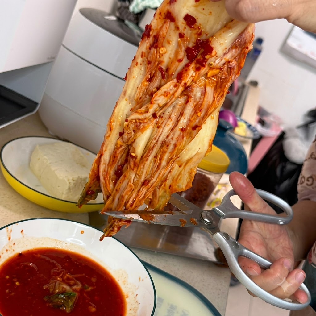 Eine silberne Küchenschere schneidet ein Stück rotes Kimchi über einer weißen Schüssel voller scharfer roter Soße.