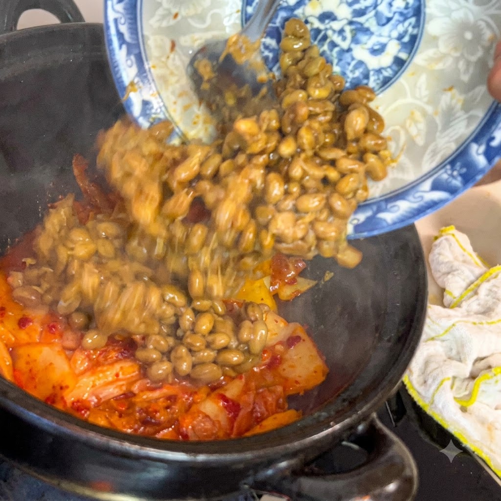 Fermentierte Sojabohnen gleiten von einem blau-weiß dekorierten Teller in einen dunklen Kochtopf voller brutzelndem Kimchi.