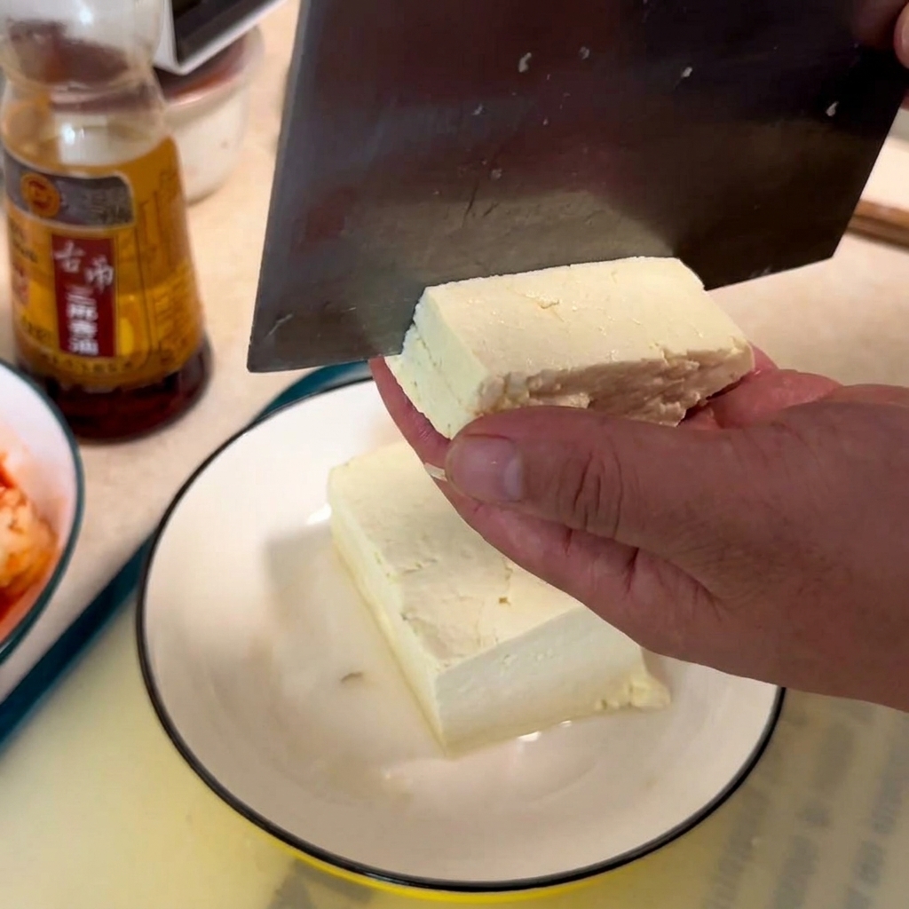 Eine Hand hält einen Block weißen Tofu, während ein Beil ihn auf einem weißen Teller in Würfel schneidet.