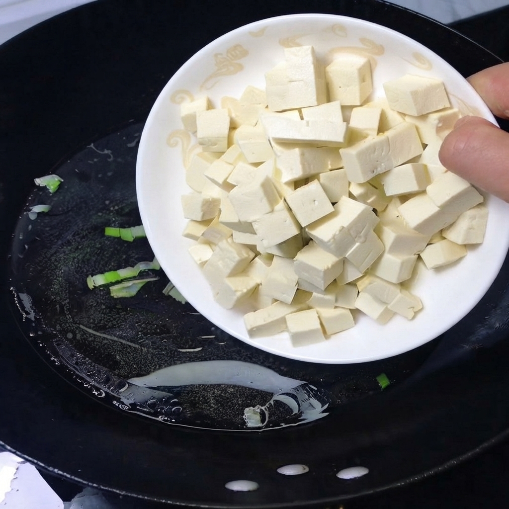 Ein Teller gewürfelter weißer Tofu wird in einen schwarzen Wok mit kochendem Wasser gegeben.