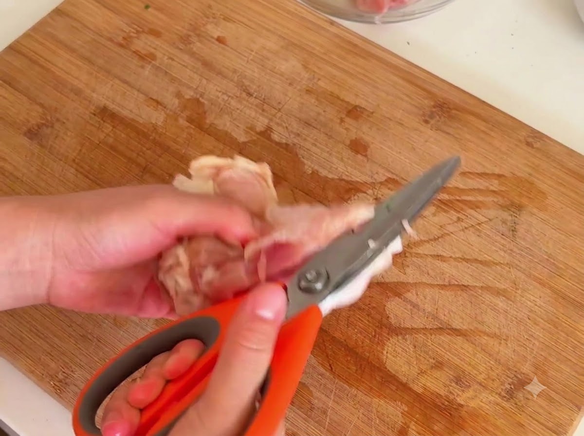 Eine Schere schneidet Fleisch vom rohen Hähnchenknochen auf einem Holzbrett.