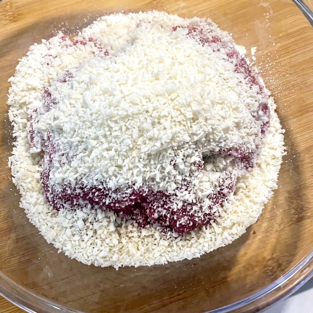 Weiße Semmelbrösel fallen aus einer Schüssel direkt auf rohes Rinderhackfleisch in einem Glasbehälter.