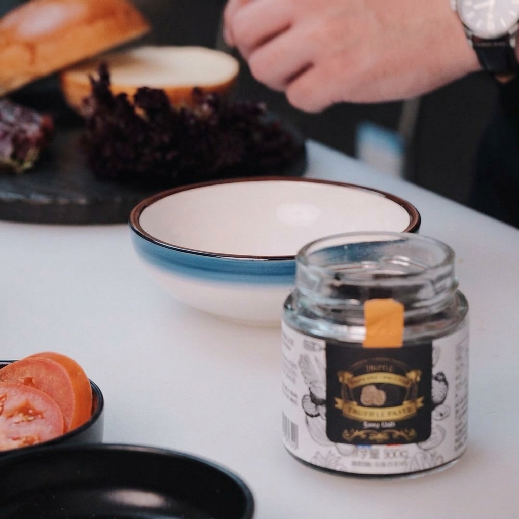 Ein kleiner Löffel schöpft weiße Mayonnaise in eine Keramikhüssel mit blauem Rand auf einer weißen Oberfläche.