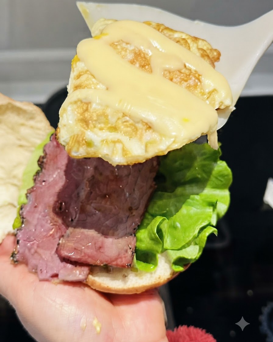 Ein Pfannenwender hebt ein mit geschmolzenem Schnittkäse belegtes Spiegelei über den Burger.