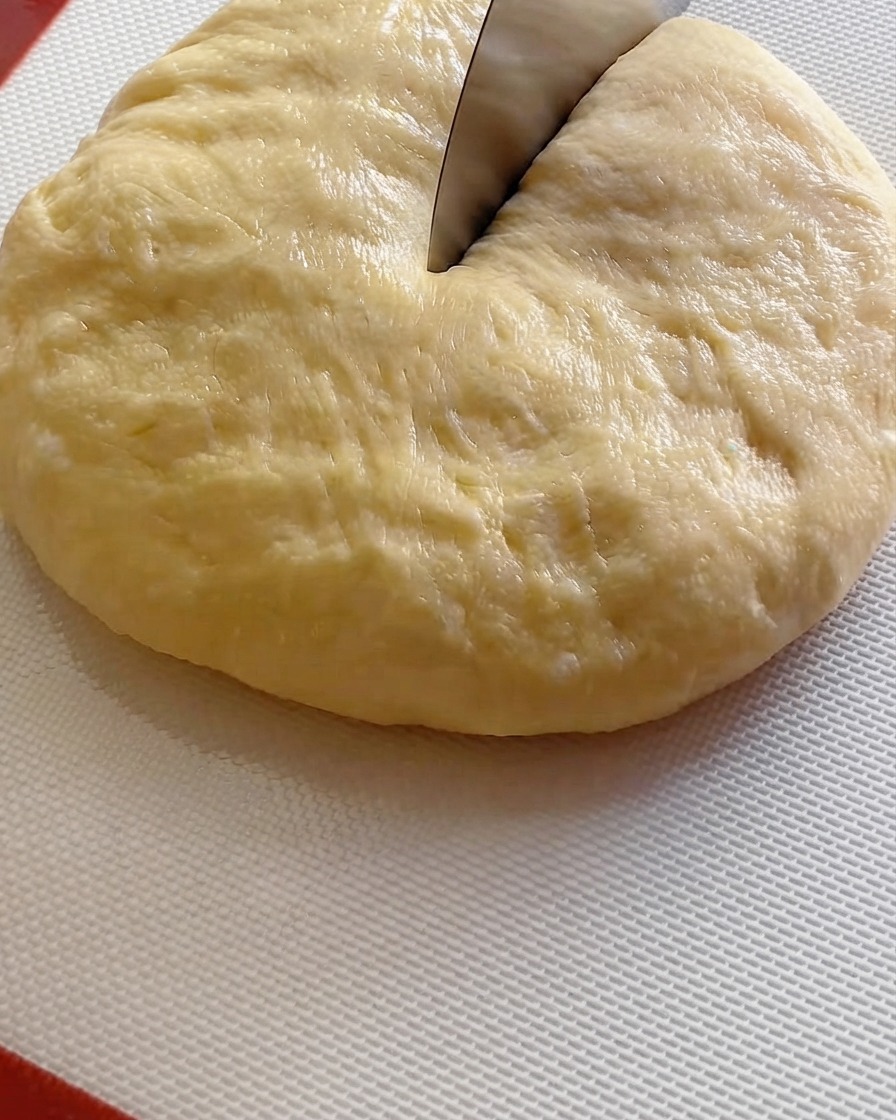 Ein Teigschaber teilt eine große Teigkugel auf einer weißen Matte in neun Portionen.