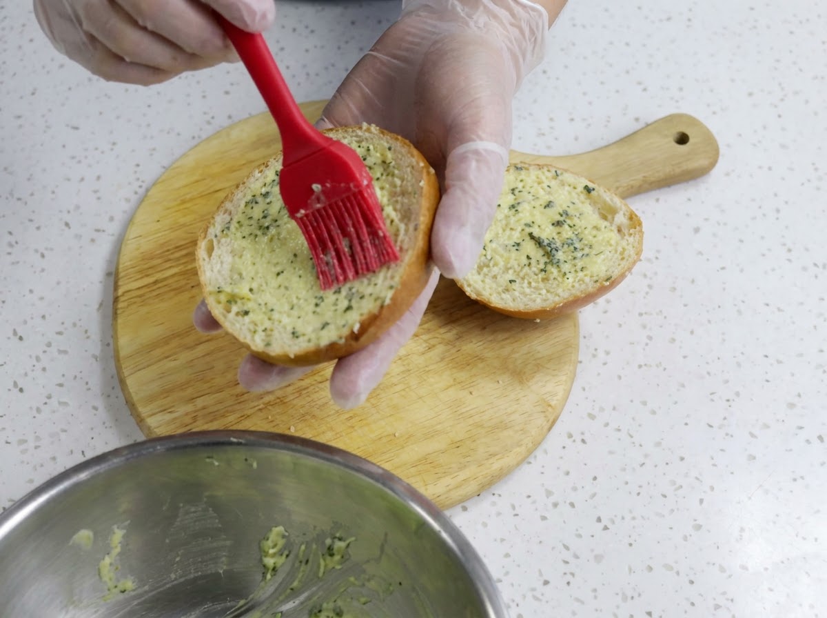 Eine Hand streicht mit einem roten Silikonpinsel Knoblauchbutter auf eine Burgerbrötchenhälfte.