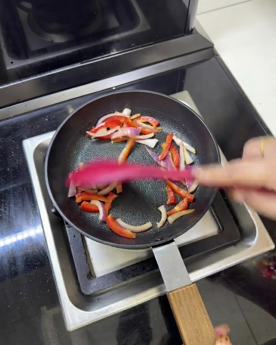 Vorgekochte Fleischstreifen werden zusammen mit roten Zwiebeln und Paprika in einer Pfanne angebraten.