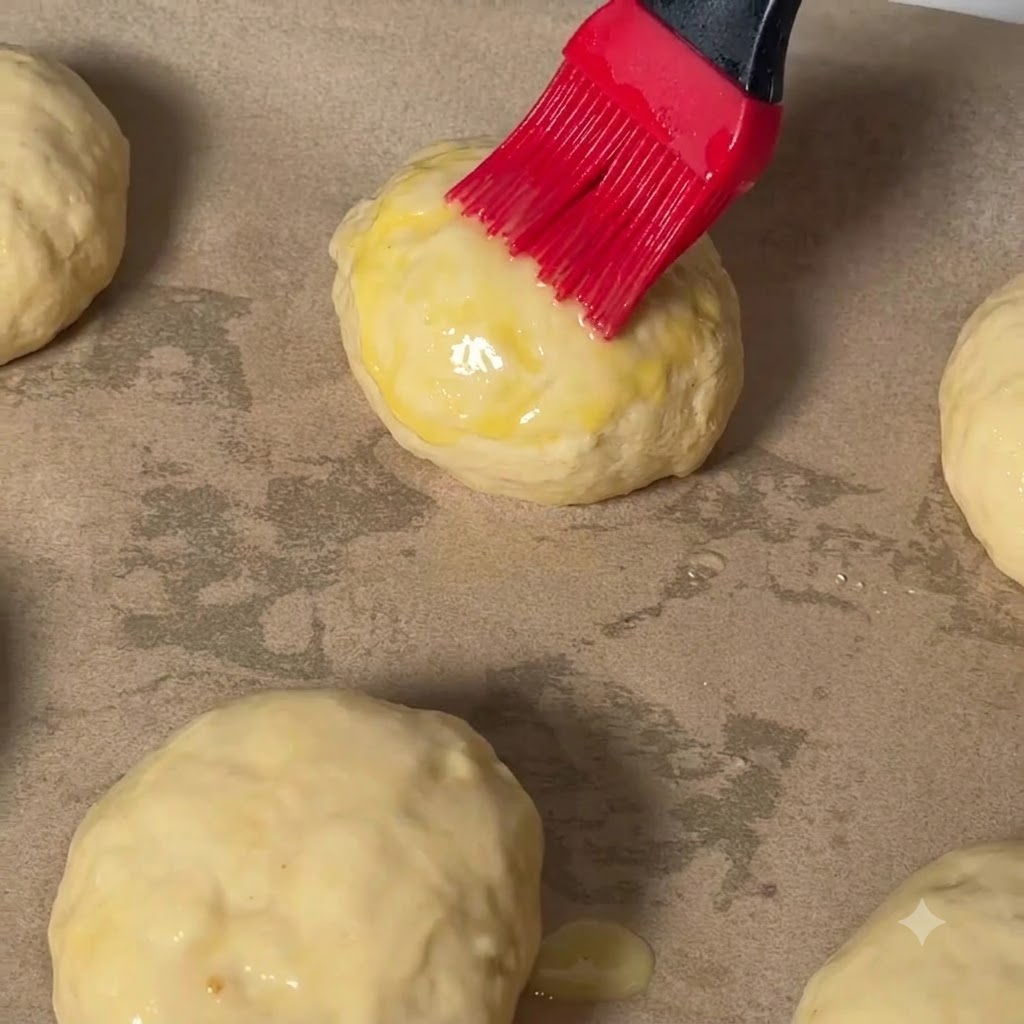 Ein roter Backpinsel trägt Eigelb auf die Oberseite einer Cheeseburger-Teigkugel auf Backpapier auf.