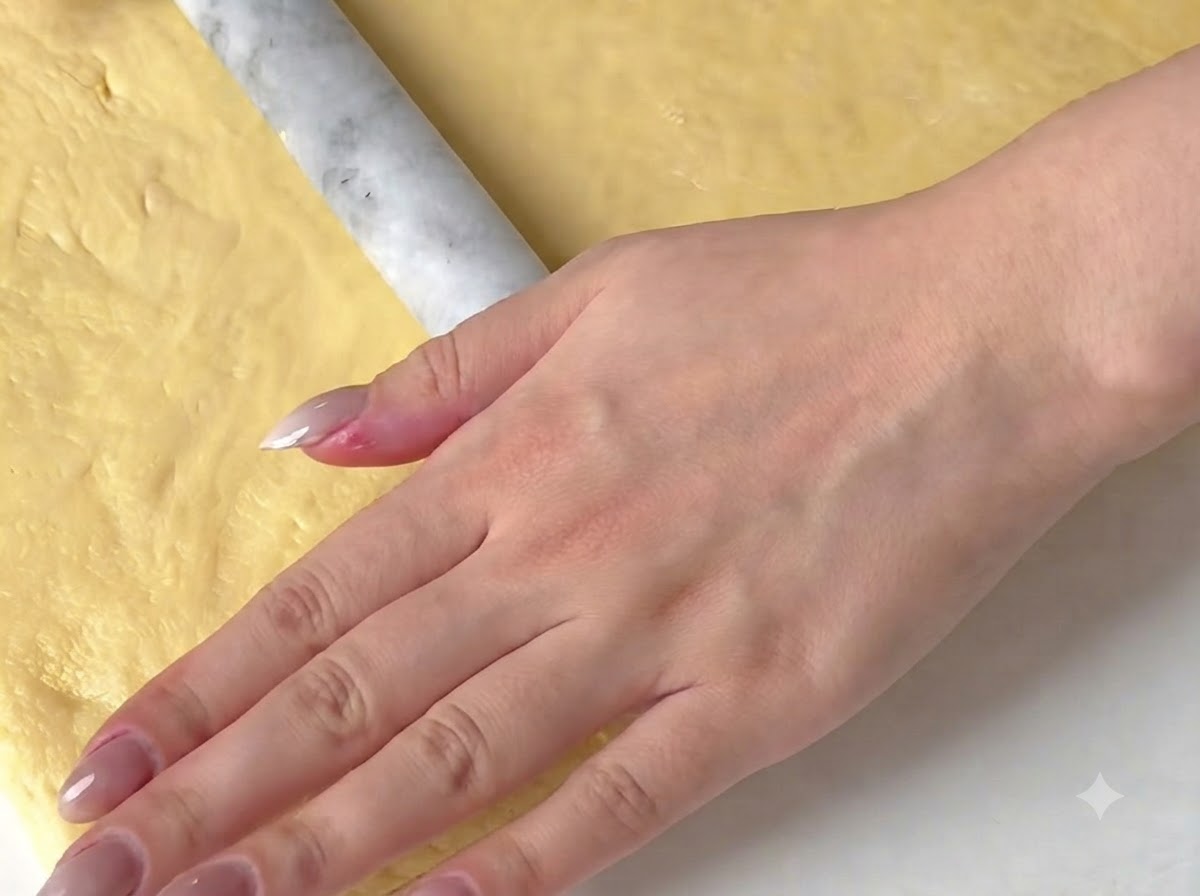Eine Hand rollt gelben Teig mit einem Teigroller auf einer Arbeitsfläche flach aus.