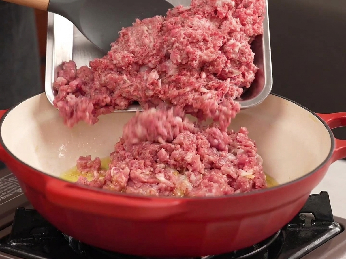 Rinder- und Schweinehackfleisch wird von einem Metalltablett in einen roten Topf mit heißem Öl gegeben.