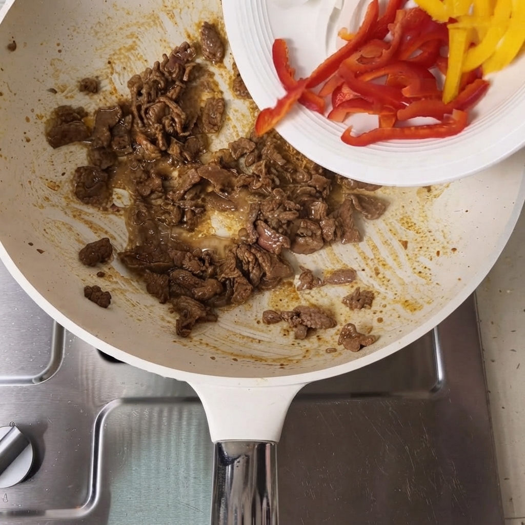 Geschnittene rote und gelbe Paprika werden von einem weißen Teller in die Pfanne mit gebratenem Rindfleisch gegeben.
