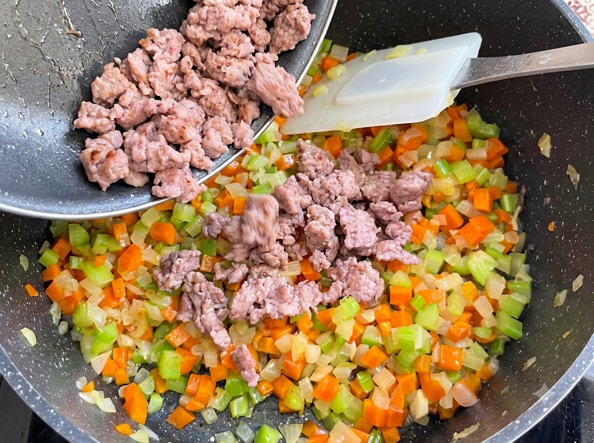 Gegartes Bratwurstbrät wird aus einer Pfanne in den Gemüsetopf gegossen und untergerührt.