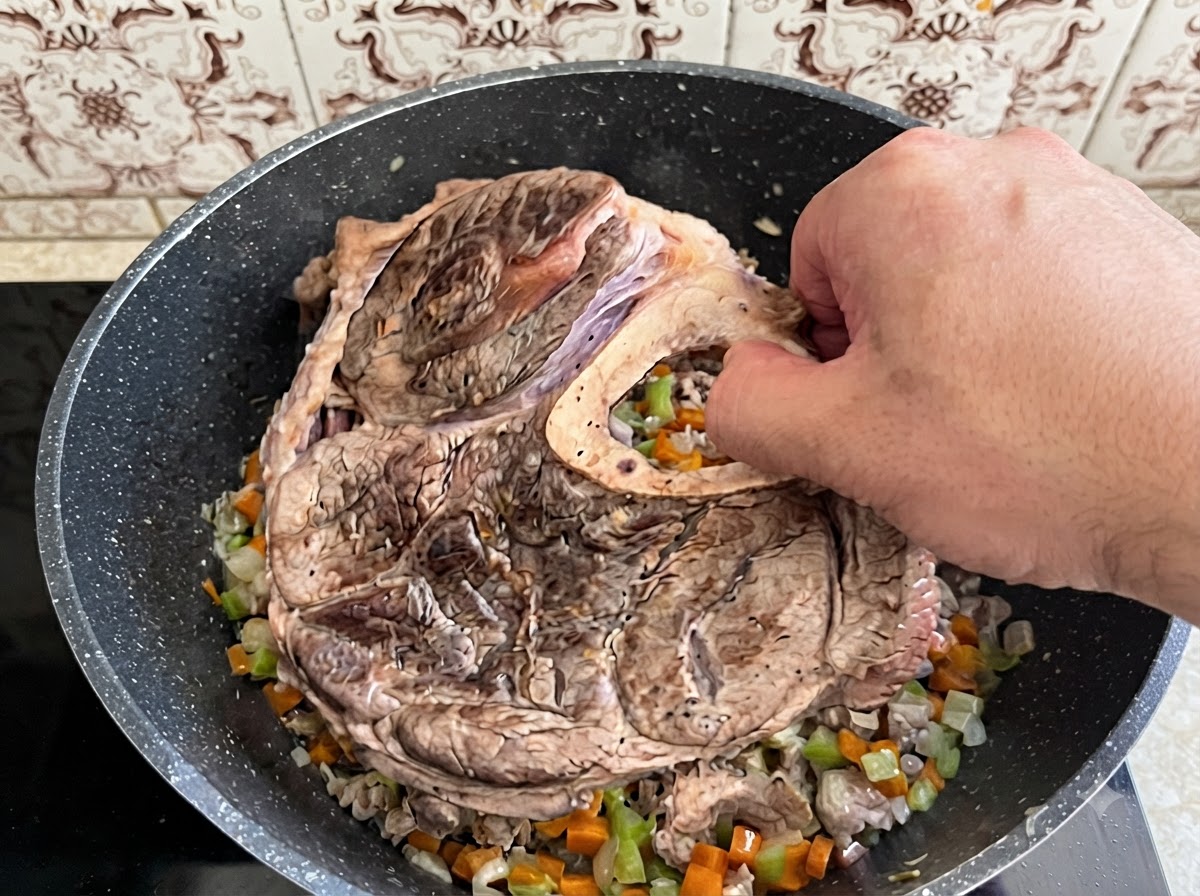 Eine Hand legt ein großes Stück angebratenes Rinderbeich und Knochen in einen mit gekochtem Gemüse gefüllten Topf.