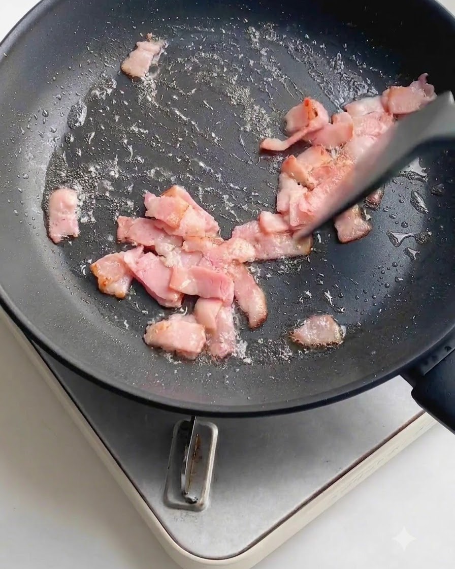 Frischer gehackter Knoblauch ist über die bratenen Speckstücke in einer dunklen Antihaft-Bratpfanne gestreut.