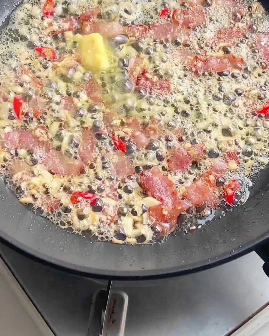 Gekochte Nudeln werden in eine Bratpfanne gegeben, die mit blubberndem Speck, geschmolzener Butter, gehacktem Knoblauch und geschnittenen roten Chilis gefüllt ist.